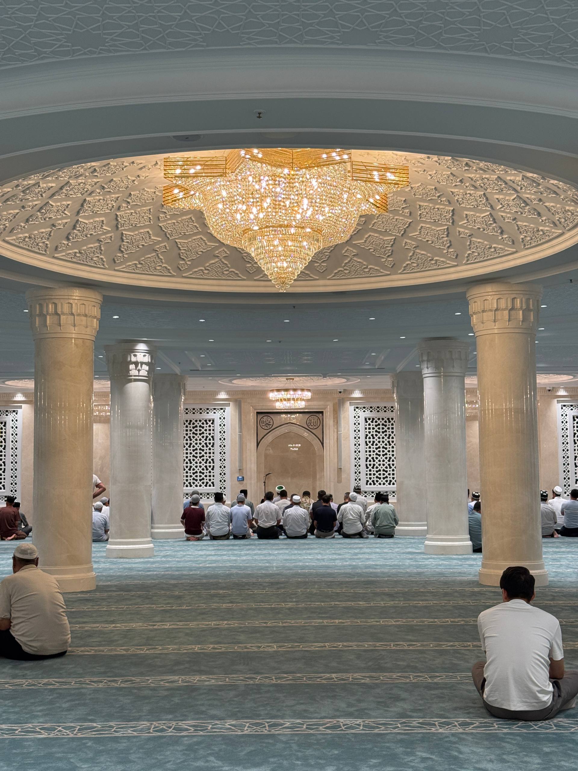 People sitting inside a mosque hall beneath a large chandelier