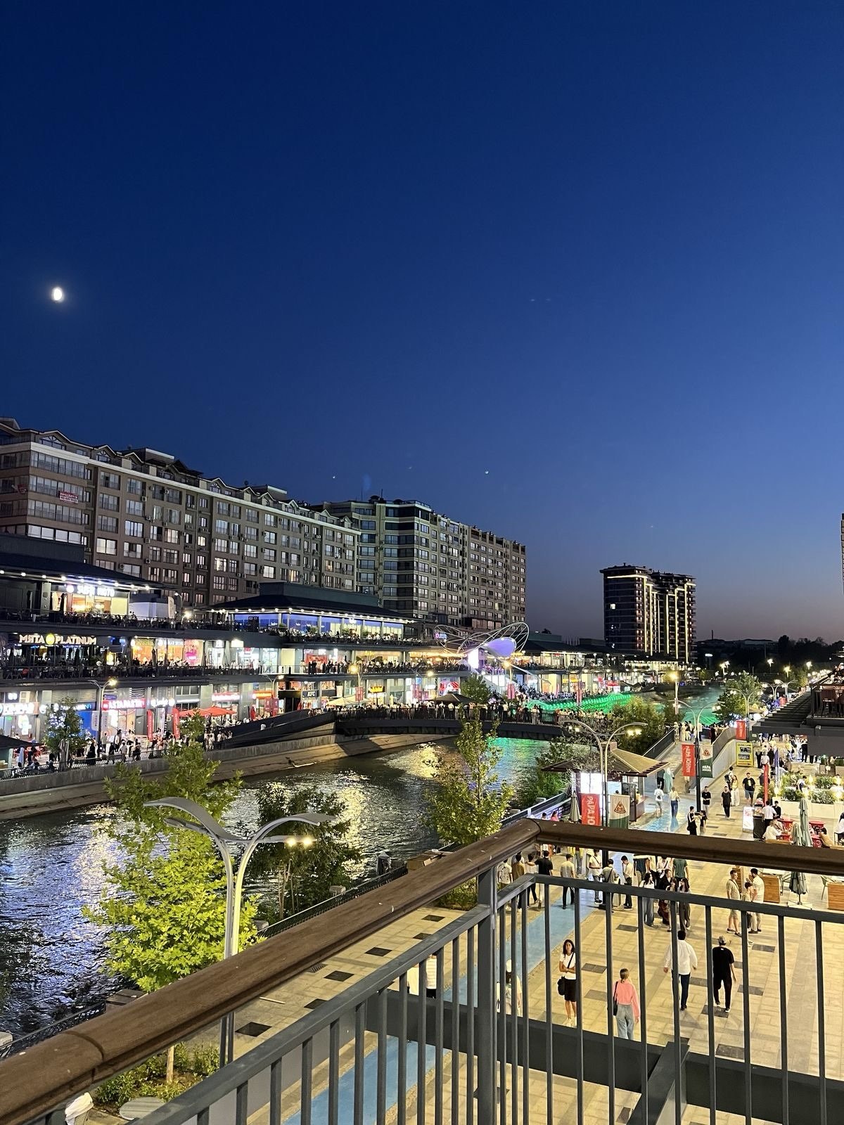 A busy canal-side promenade at dusk with the moon visible in the sky